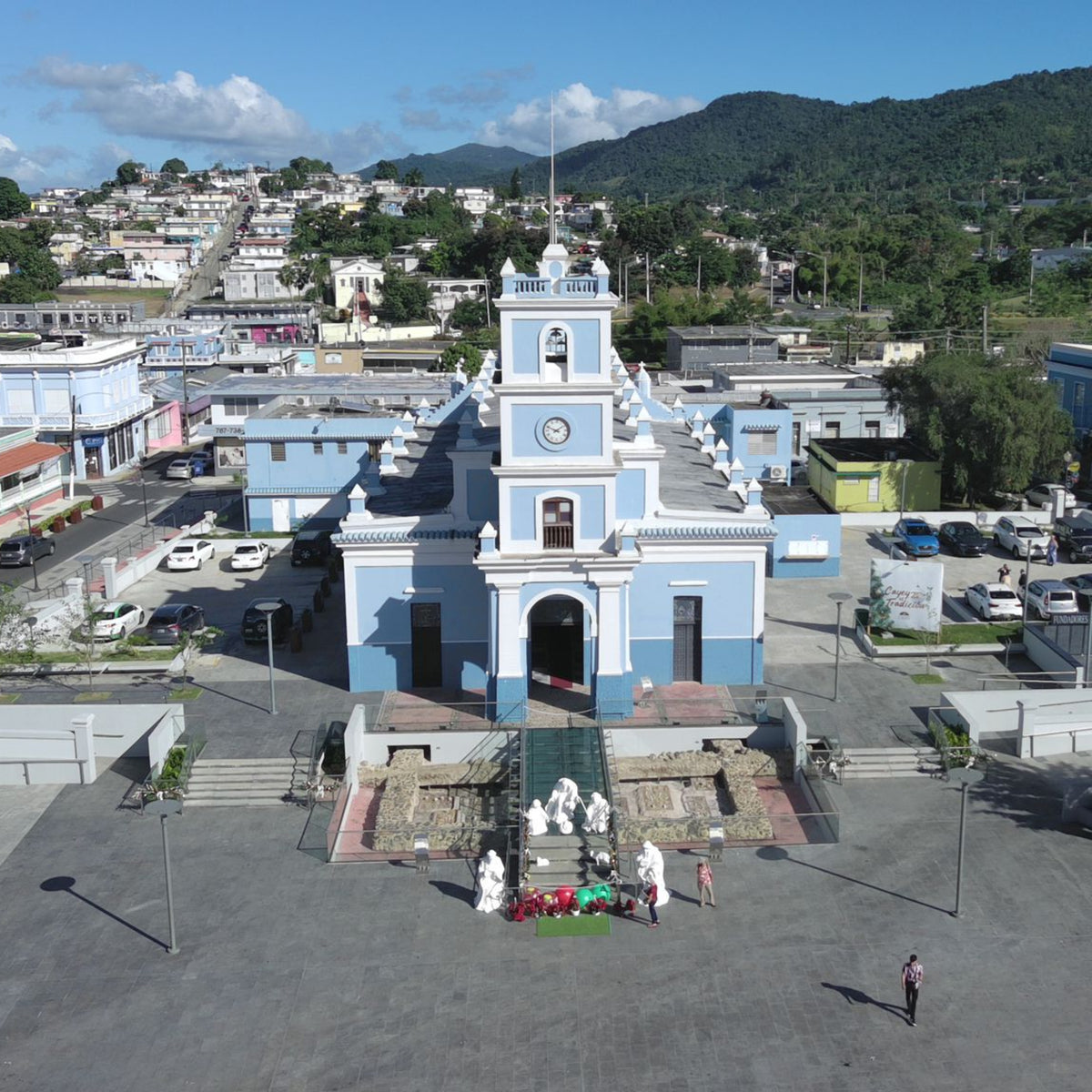 Aerial view of a town square with a prominent blue and white building, likely a church, surrounded by other buildings and greenery.